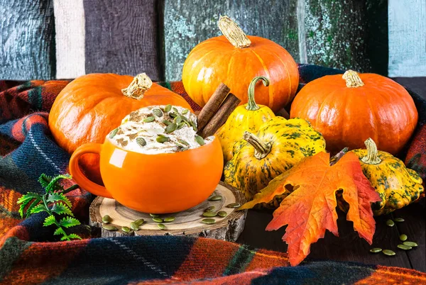 Pumpkin latte with cream, seeds, spices in pumpkin orange cup, autumn leaves, pumpkins, woolen scarf on wooden background.
