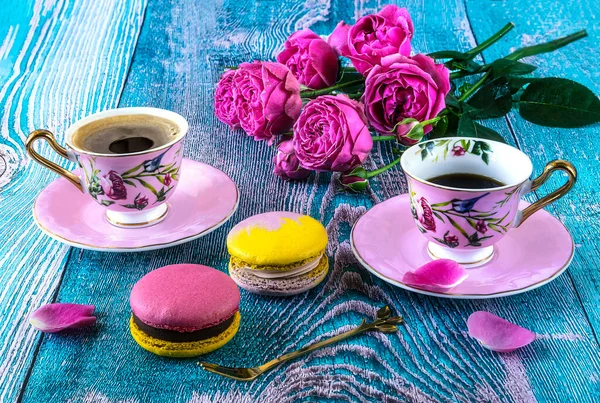 Macaroons and two pink cups of coffee and peony blooming pink roses, composition on a blue background