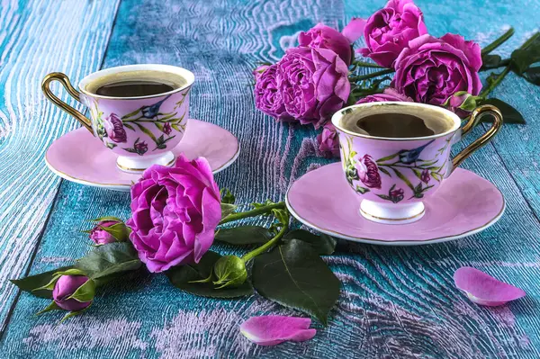 Two pink cups of coffee and peony blooming pink roses, composition on a blue background