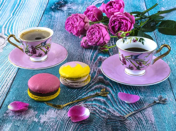 Macaroons and two pink cups of coffee and peony blooming pink roses, composition on a blue background