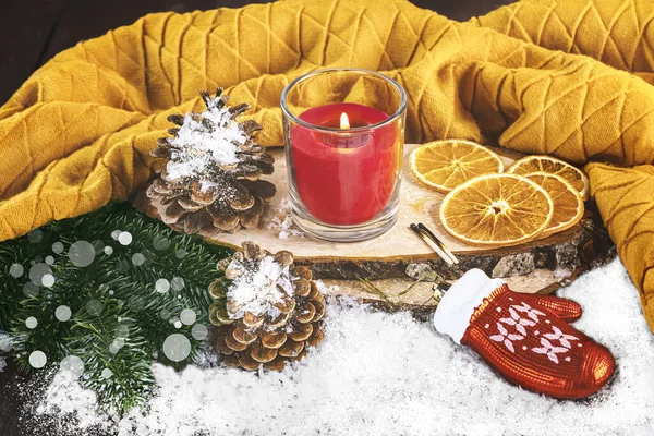 Winter cozy still life with red burning candle, Christmas tree toy glove, snowy cones, dried oranges, yellow scarf.