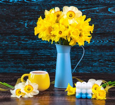 Mavi bir vazoda bir buket nergis çiçeği, sarı bir fincan kahve, koyu ahşap zemin üzerinde mavi bir mum. Sarı çiçeklerle dolu bahar