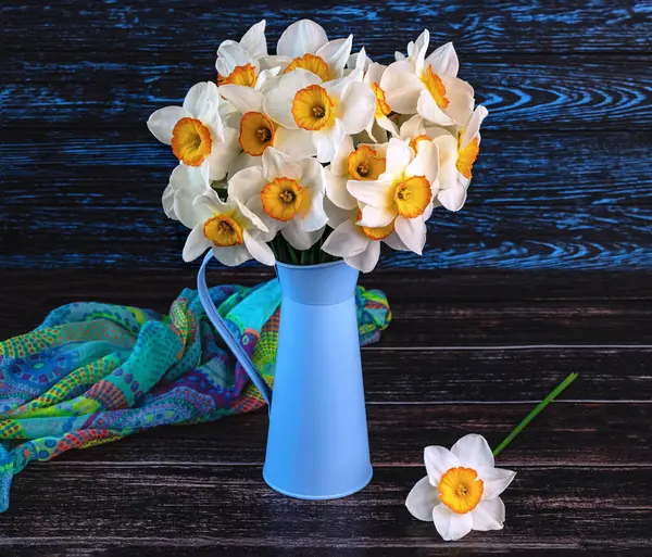 Bouquet of daffodils flowers in blue vase, colorful shawl on dark wooden background. Spring still life with yellow flowers