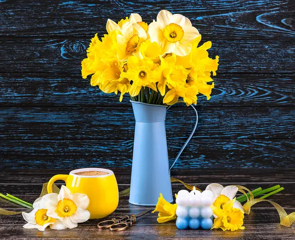 A bouquet of daffodil flowers in a blue vase, a yellow cup of coffee, a blue candle on a dark wooden background. Spring still life with yellow flowers