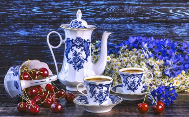 Kirazlı klasik bir sepet, iki fincan kahve, mavi ve beyaz renkli bir kahve demliği, bir buket kır çiçeği. Yaz hala kirazlarla dolu