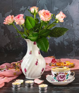 Güzel bir vazoda bir buket pembe gül, bir fincan kahve, bir şal, koyu arkaplanda mumlar ve yapraklar..