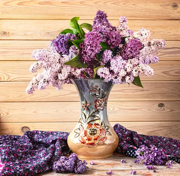 Güzel bir demet leylak çiçeği yüksek kaliteli bir vazoda, açık ahşap bir arka planda mor bir şal. Bahar, leylak dallarıyla hayat dolu.