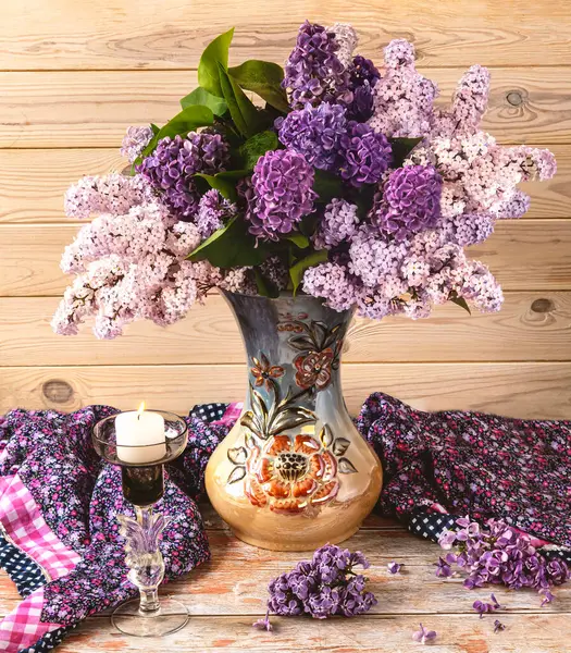  Güzel bir demet leylak çiçeği uzun bir vazoda, bir şamdan, açık ahşap bir arka planda mor bir şal..