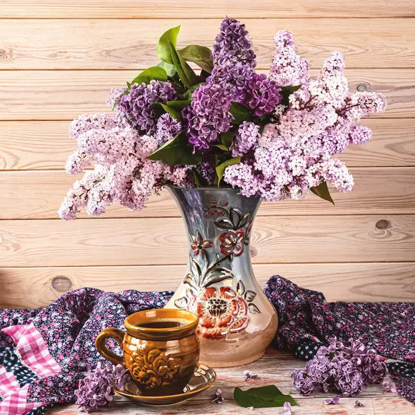 Güzel bir demet leylak çiçeği uzun bir vazoda, bir fincan kahve, açık ahşap arka planda mor bir şal..