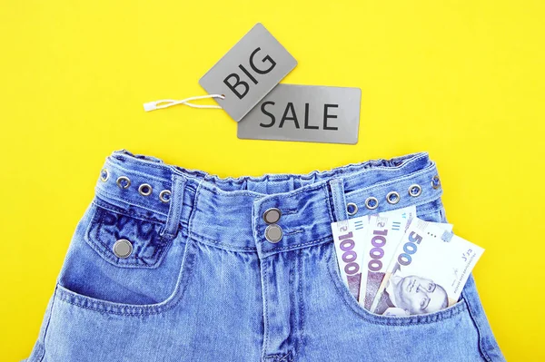 Flat lay, detail of denim jeans with Ukrainian hryvnia bills in pocket with grey label on yellow background. Shopping and discount concept. Black Friday.