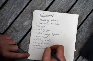 gril writing a camping list in a piece of paper. writing a check list with a pencil and a phone