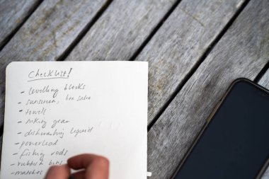 gril writing a camping list in a piece of paper. writing a check list with a pencil and a phone