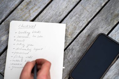 gril writing a camping list in a piece of paper. writing a check list with a pencil and a phone