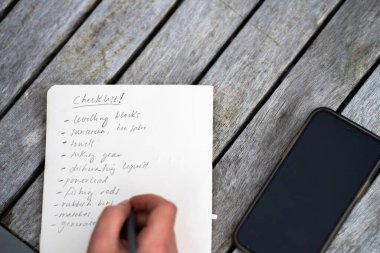 gril writing a camping list in a piece of paper. writing a check list with a pencil and a phone