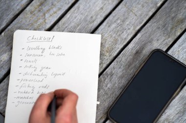 gril writing a camping list in a piece of paper. writing a check list with a pencil and a phone