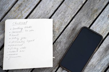 gril writing a camping list in a piece of paper. writing a check list with a pencil and a phone