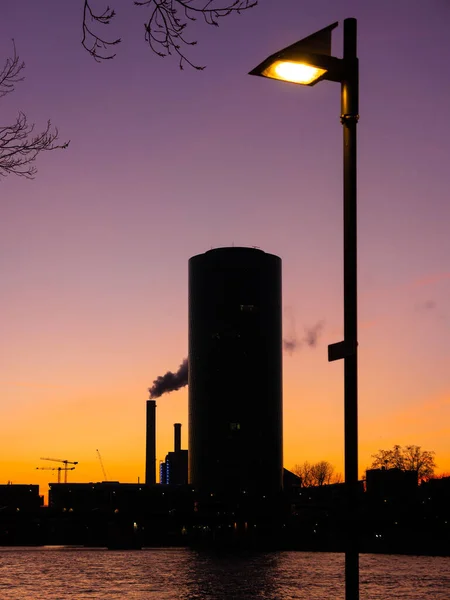 Street lamp and a silhouette of a round skyscraper and industrial buildings along the banks of the Main River in Frankfurt, Germany at sunset