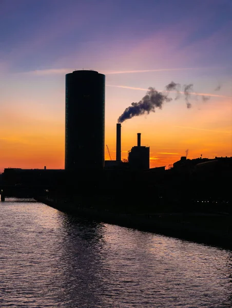 Silhouettes of a round skyscraper and an industrial buildings along the banks of the Main River in Frankfurt, Germany at sunset
