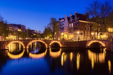Amsterdam 'da sakin bir akşam, alacakaranlığın yumuşak parıltısıyla aydınlandı. Güzel bir kemer köprüsü büyüleyici mimari ve nehir manzarasının ortasındaki bir kanala yansıyor..