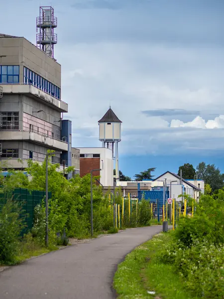 stock image Urban industrial scene with modern building, water tower, greenery, path, yellow poles, and cloudy sky.