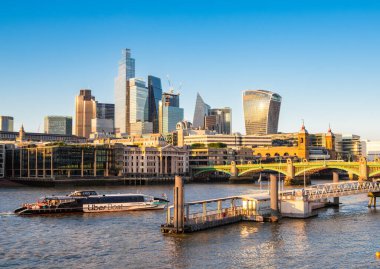 Londra, Birleşik Krallık - 25 Haziran 2024: Thames nehri boyunca modern gökdelenlerin yer aldığı canlı bir şehir manzarası, günbatımının sıcak parıltısını yansıtıyor. Uber Bot geçiyor..