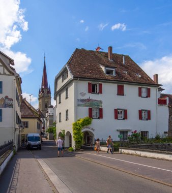 Radolfzell, Almanya - 2 Ağustos 2024: Alman kasabası Radolfzell 'de pitoresk bir sokak sahnesi, kırmızı panjurlu beyaz bir bina, arka planda saat kulesi ve yol boyunca yürüyen insanlar.