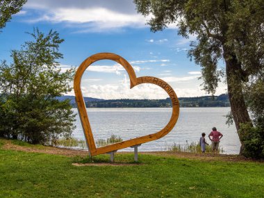 Radolfzell, Almanya - 2 Ağustos 2024: Büyük ahşap bir kalp heykeli Constance Gölü 'nün kenarında duruyor, arka planda iki kişi manzaranın keyfini çıkarıyor.