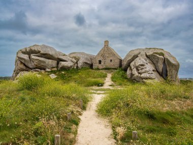 Kerlouan 'da iki büyük kaya arasına yerleştirilmiş küçük bir taş ev, bulutlu bir gökyüzünün altındaki çimenli bir arazide..