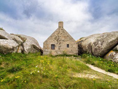 Kerlouan 'da iki büyük kaya arasına yerleştirilmiş küçük bir taş ev, bulutlu bir gökyüzünün altındaki çimenli bir arazide..