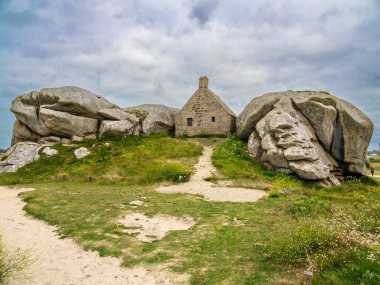 Kerlouan 'da iki büyük kaya arasına yerleştirilmiş küçük bir taş ev, bulutlu bir gökyüzünün altındaki çimenli bir arazide..