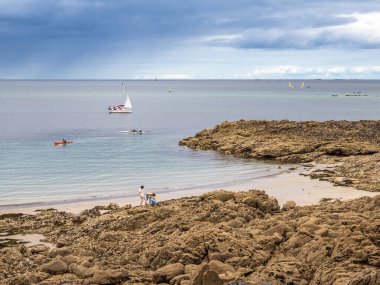 Dinard, France - July 20, 2024: Coastal scene in Brittany with rocky shores, calm sea, and a few sailboats in the distance. A person walks along the beach, and a kayak is visible on the water. clipart