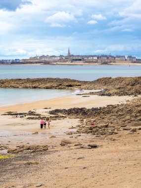 Dinard, France - July 20, 2024: A scenic view of a rocky beach with people walking, overlooking a historic coastal town of Saint Malo in the background under a cloudy sky. clipart