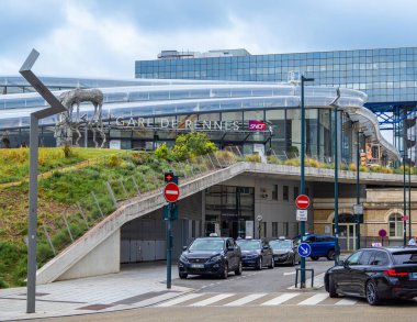 Rennes, Fransa - 26 Temmuz 2024: Ön planda park halindeki arabalar ve trafik işaretlerinin yer aldığı cam cephe ve metal heykellerle modern tren istasyonu girişi.
