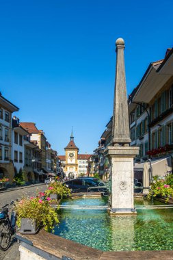 Murten, İsviçre - 21 Eylül 2024: Büyüleyici bir İsviçre meydanındaki taş çeşme, tarihi binalar ve açık mavi gökyüzünün altında bir saat kulesi.