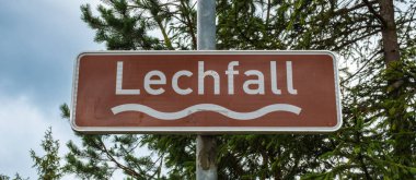 Fussen, Bavyera 'daki Kahverengi Lechfall Şelalesi yol tabelası bulutlu bir gökyüzünün altındaki yeşil ağaçların arasında.