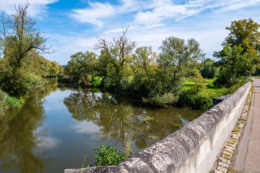 Wornitz river scene in Ebermergen with a bridge, lush greenery on both banks, reflecting the blue sky and clouds. Trees with varying foliage surround the water, creating a peaceful natural landscape. clipart