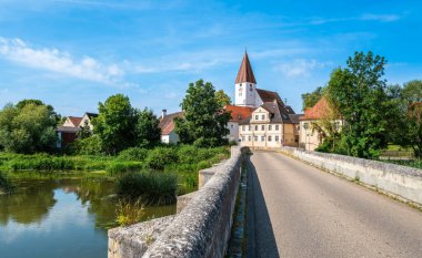 A scenic view of a quaint village of Ebermergen surrounded by lush greenery and a calm river. The image captures a stone bridge leading into the village under a clear blue sky. clipart