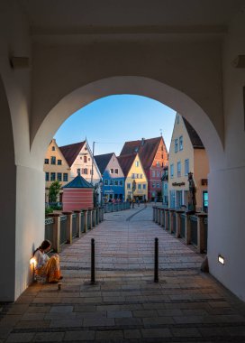 Donauworth, Germany - August 6, 2024: View through archway of Rieder Tor gate leading to a charming town square. Colorful buildings line the square, a person sitting against the wall clipart