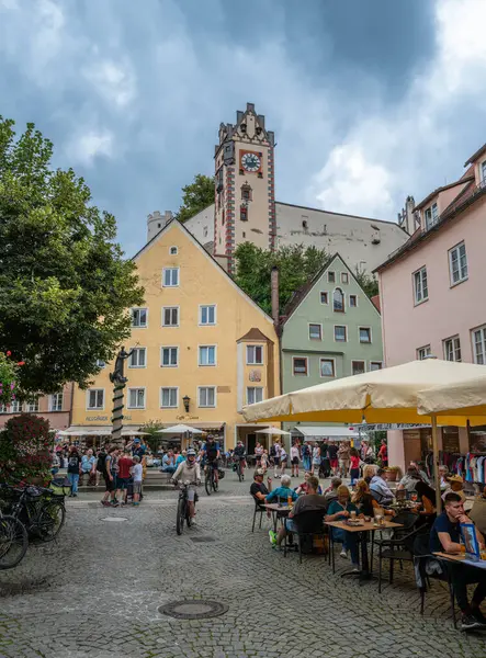 Fussen, Almanya - 5 Ağustos 2024 Fussen 'de renkli binaları, açık kafeleri ve bulutlu gökyüzünün altında tarihi bir saat kulesi olan canlı bir Alman kasaba meydanı..