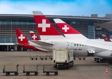 Zürih, İsviçre - 13 Kasım 2024: Zürih havaalanına park etmiş bir dizi İsviçre havayolu uçağı. Zemin servis araçları ve bagaj arabaları ön planda görünür..