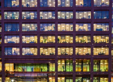 Londra, Birleşik Krallık - 27 Ekim 2024: Günbatımında Canary Wharf 'taki modern bir ofis binası cephesi, çeşitli ofis ayarları ve bitkilerle aydınlatılmış pencereler sergileniyor.