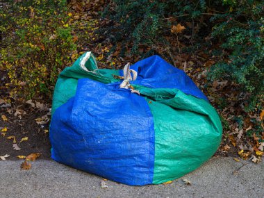 Yapraklarla dolu büyük mavi ve yeşil bir tente torbası çimentoyla çevrili beton bir yolda oturur..