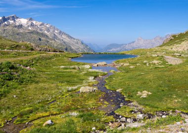 İsviçre 'de Sustenpass' ın sakin bir manzarası. Arka planda yemyeşil çayırlar ve kayalık dağlarla çevrili berrak mavi bir göl var..