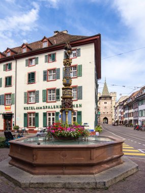 Basel, İsviçre - 17 Ağustos 2024: Ön planda renkli çiçekler bulunan tarihi bir çeşmenin yer aldığı sokak manzarası, geleneksel binalar ve bir ortaçağ kapısı Spalentor.