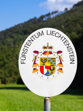 Balzers, Lihtenştayn - 16 Ağustos 2024: Liechtenstein Prensliği armasını gösteren sınır işareti, arka planda tepeler olan yeşil bir manzaraya karşı kurulmuş.
