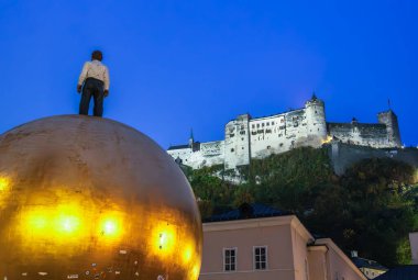 Salzburg, Avusturya - 24 Ekim 2024: Salzburg, Kapitelplatz 'daki Sphaera heykeli - altın bir kürenin üzerinde rahatlatıcı bir duruş içinde duran bir adam figürü. Arka plandaki şato.