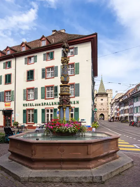 Basel, İsviçre - 17 Ağustos 2024: Ön planda renkli çiçekler bulunan tarihi bir çeşmenin yer aldığı sokak manzarası, geleneksel binalar ve bir ortaçağ kapısı Spalentor.