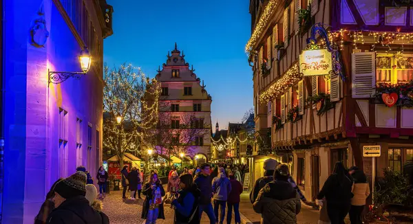 Colmar, Fransa - 28 Kasım 2024: Colmar, Alsace 'de insanların yürüdüğü ve binaların Noel ışıkları ve süslemeleriyle süslendiği şenlikli bir sokak sahnesi.