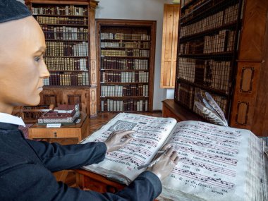 Michaelbeuern, Avusturya - 22 Ekim 2024: Dini kıyafetlerle kutsanmış bir keşişi temsil eden bir manken büyük pencereli bir odada ahşap bir kürsüde duruyor, bir kitapta müzik yazıyor..