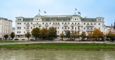 Salzburg, Avusturya - 24 Ekim 2024: Salzach Nehri kıyısında Sacher Salzburg Oteli, sonbahar ağaçları ve bulutlu gökyüzü.